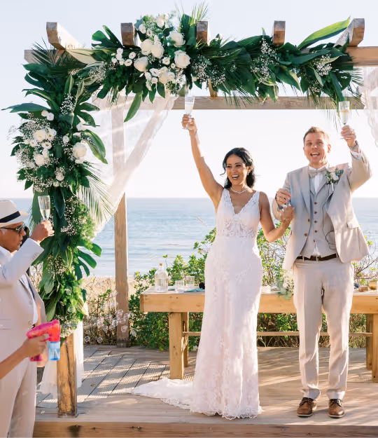 Bride and groom in wedding attire raising champagne glasses under a floral arch by the seaside.