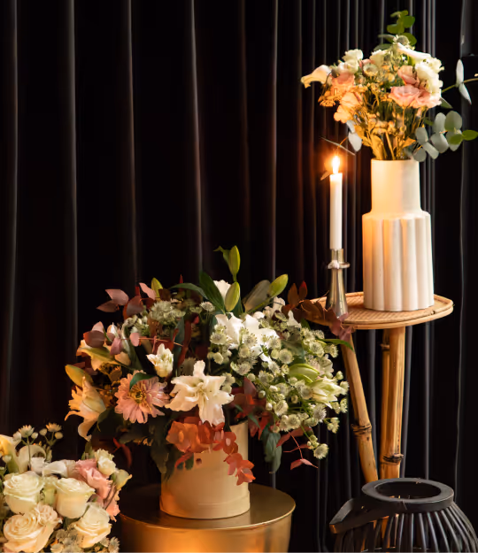 Elegant floral arrangements in vases with a lit white candle on a small round wooden table against a dark curtain background.