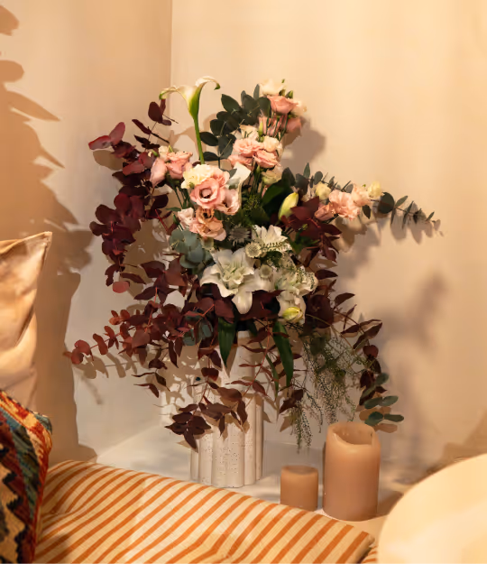 Elegant floral arrangement with pink and white flowers and dark red foliage in a white vase, beside beige candles on a table.