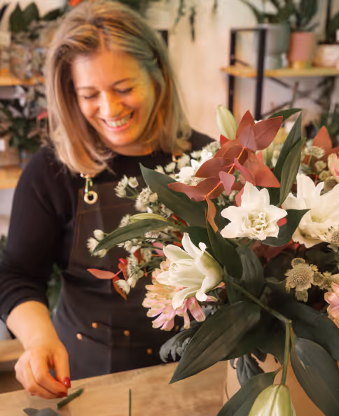 Smiling woman arranging a bouquet of white and pink flowers with green and red leaves indoors.
