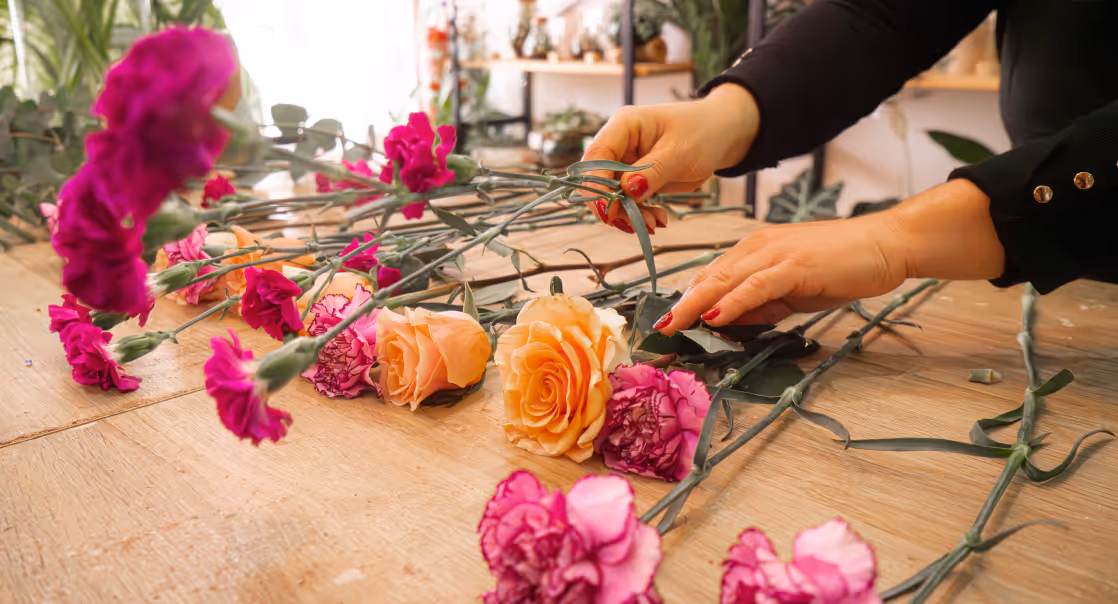 Person arranging orange roses and pink carnations on a wooden table in a flower shop.