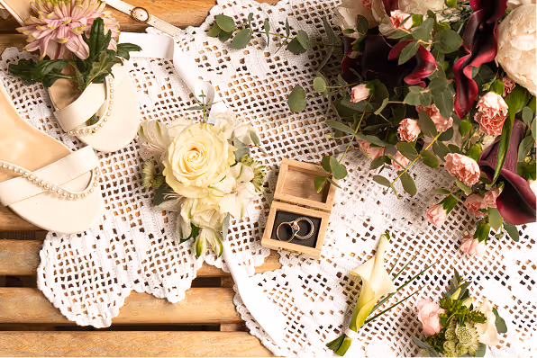 Wedding rings in a wooden box surrounded by wedding flowers, pearl sandals, and lace fabric on a wooden surface.