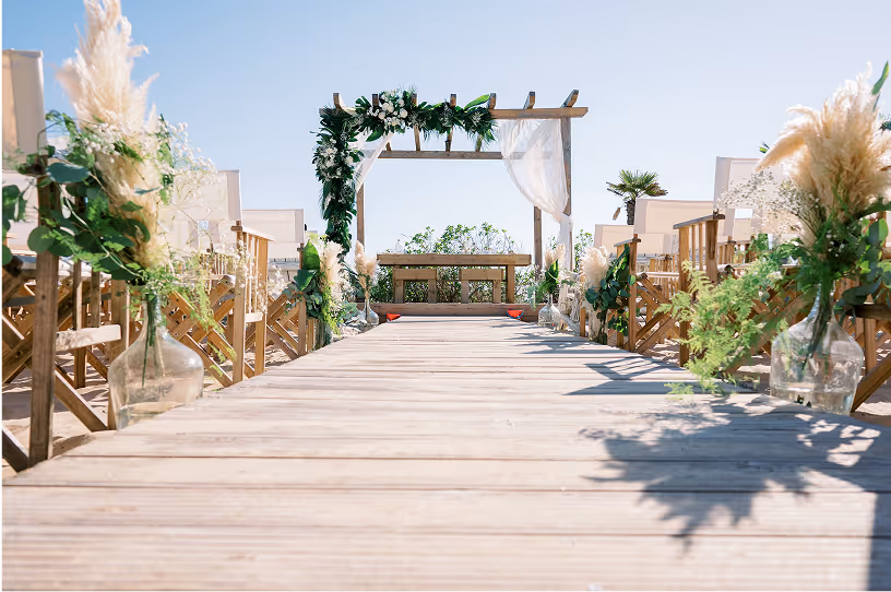 Outdoor wooden wedding aisle with flower arrangements leading to a decorated wooden arch under a clear sky.