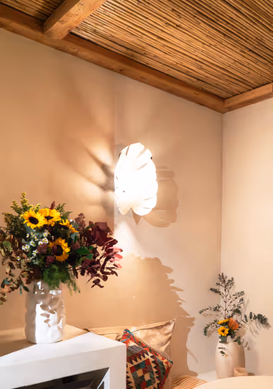 Cozy corner with a rustic wooden ceiling, lit wall sconce casting shadows, and vases holding sunflowers and greenery.