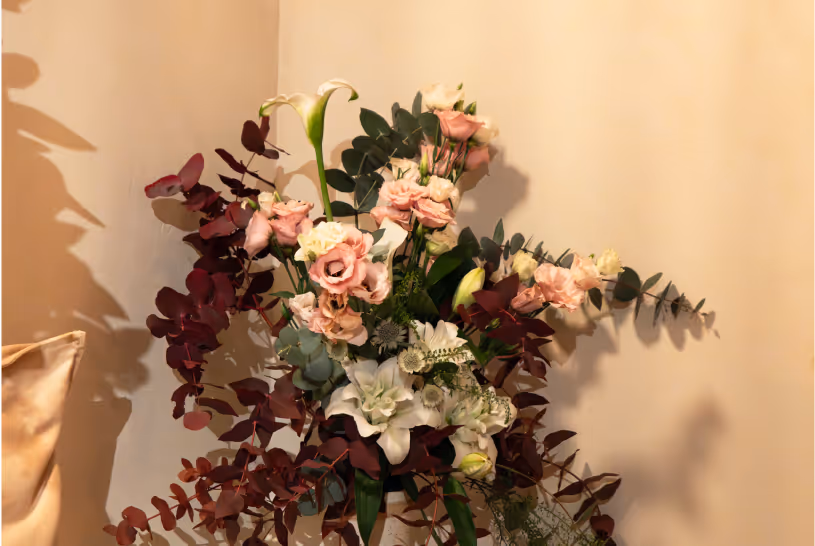 Bouquet of mixed flowers including soft pink roses, white lilies, and green foliage against a beige wall.