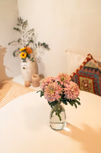 Pink chrysanthemum flowers in a small glass vase on a white table, with a blurred background featuring a patterned pillow and another vase with various flowers.