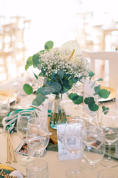Table centerpiece with white and green floral arrangement surrounded by empty wine glasses at a bright event setting.