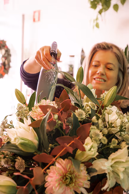 Woman watering a lush bouquet of mixed flowers and greenery indoors.