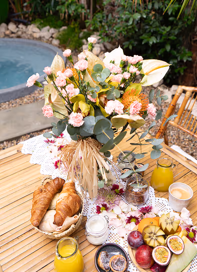 Table with a floral centerpiece, basket of croissants, jars of yogurt or milk, fresh fruits, orange juice, and a cup of coffee beside a pool.