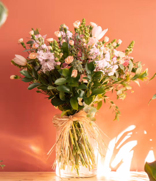Bouquet of pink and peach flowers with green foliage tied with raffia in a clear glass vase on a wooden surface against a coral background.