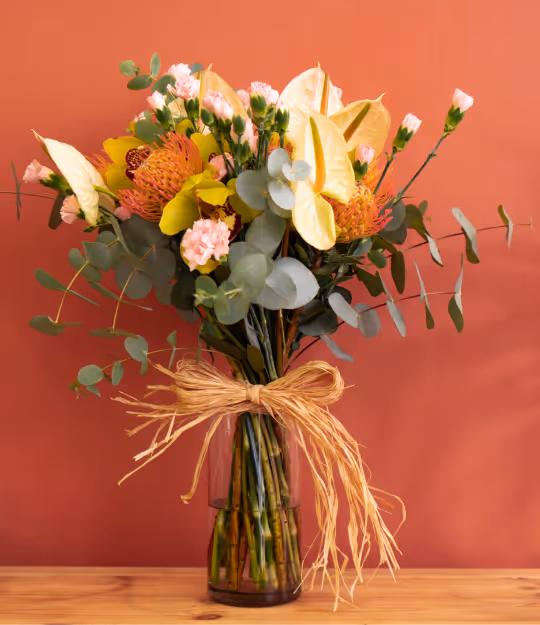 Colorful bouquet with yellow, pink, and orange flowers and green leaves in a glass vase tied with a natural twine bow on a wooden surface.