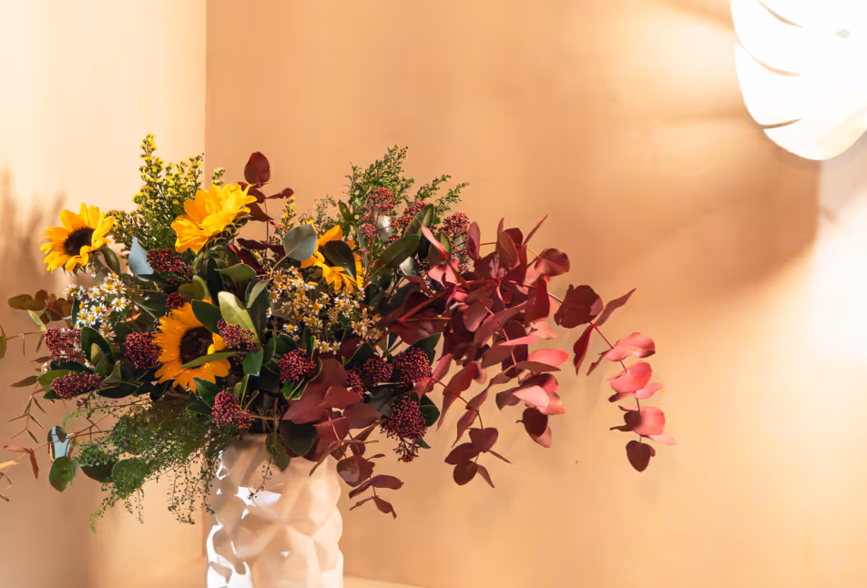White vase with large floral arrangement including sunflowers, burgundy leaves, and small white flowers against a beige wall.