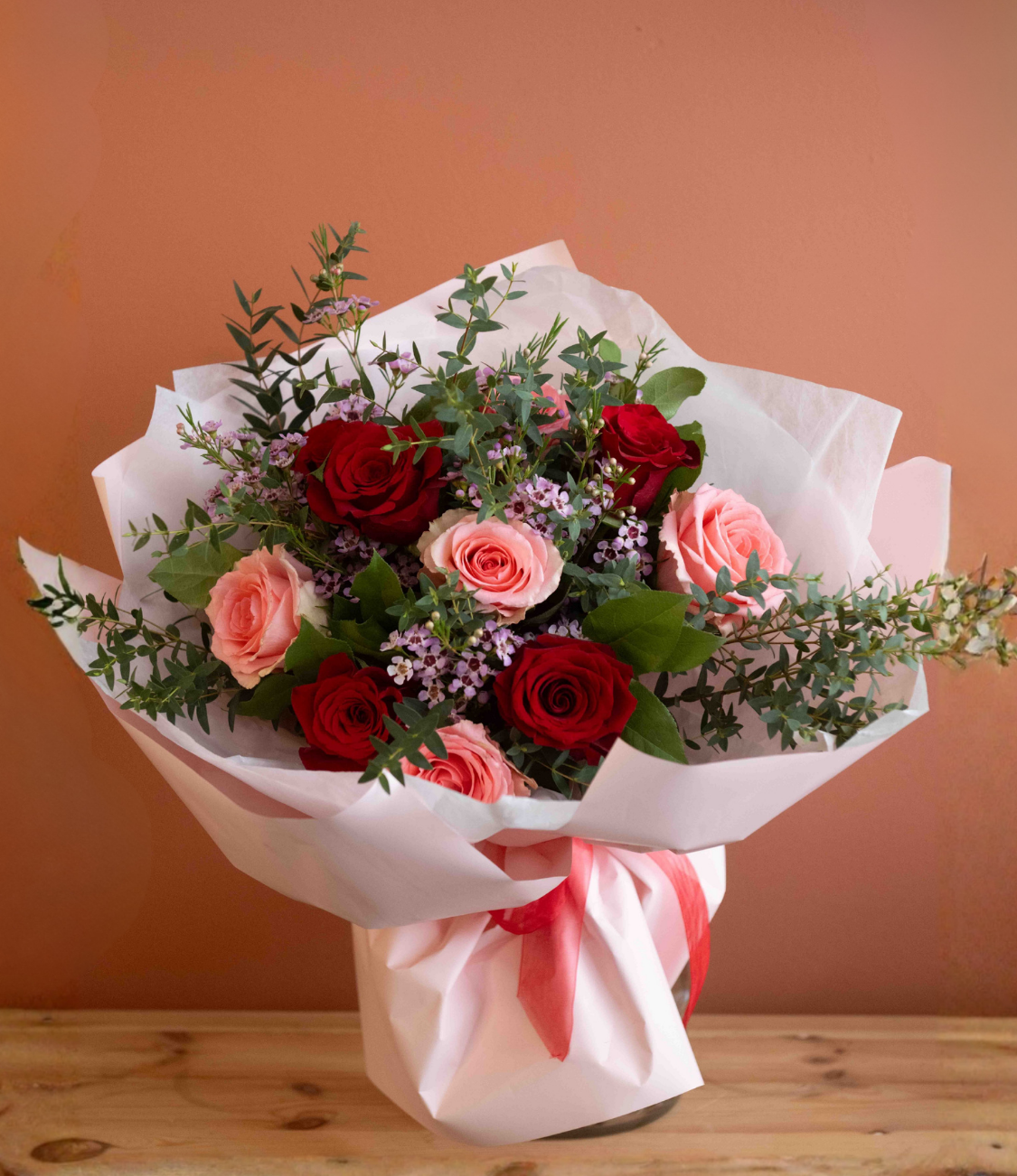 Bouquet of red roses with green foliage in a clear glass vase tied with a raffia bow on a wooden surface against a terracotta background.