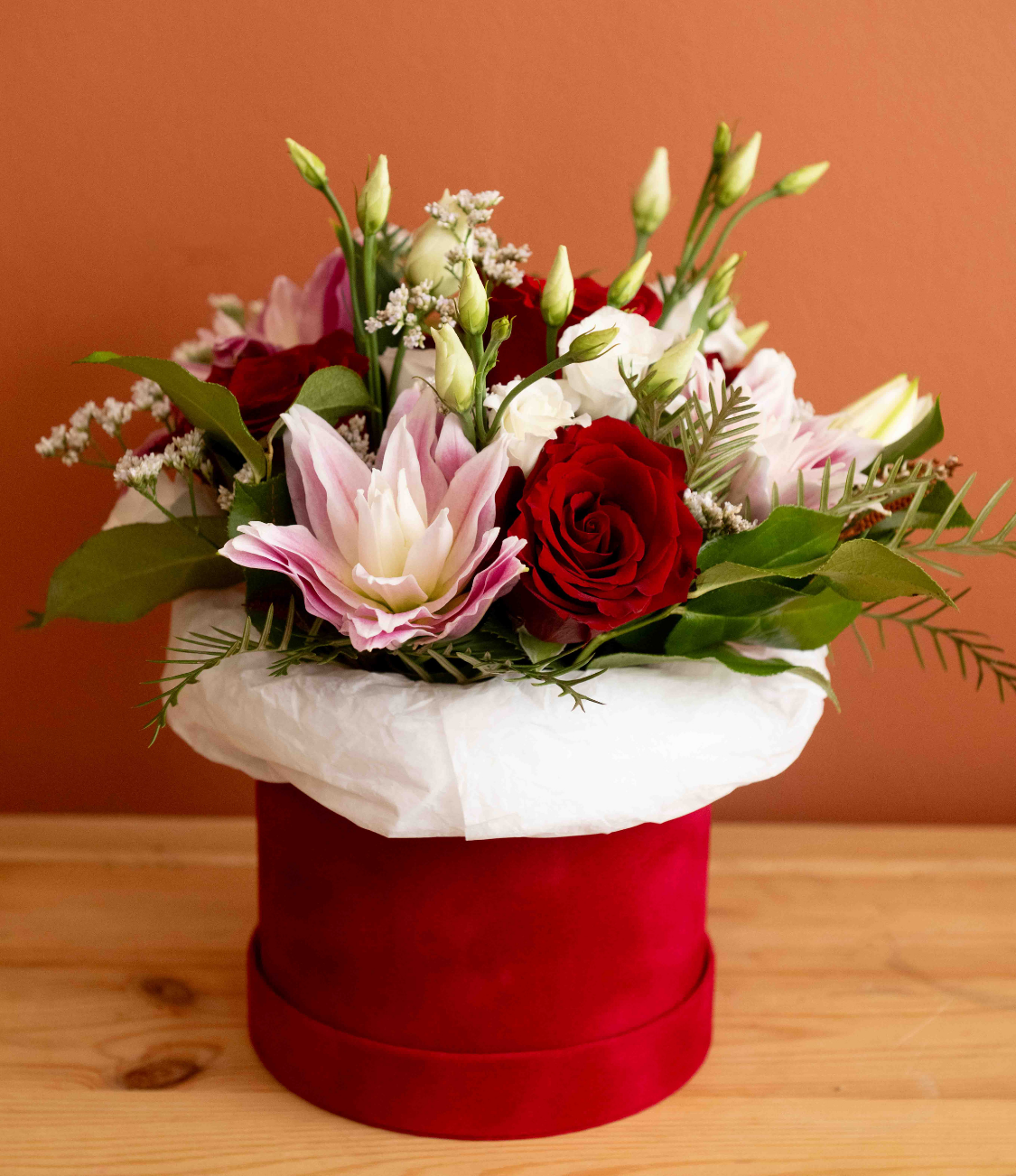Bouquet of red roses with green foliage in a clear glass vase tied with a raffia bow on a wooden surface against a terracotta background.
