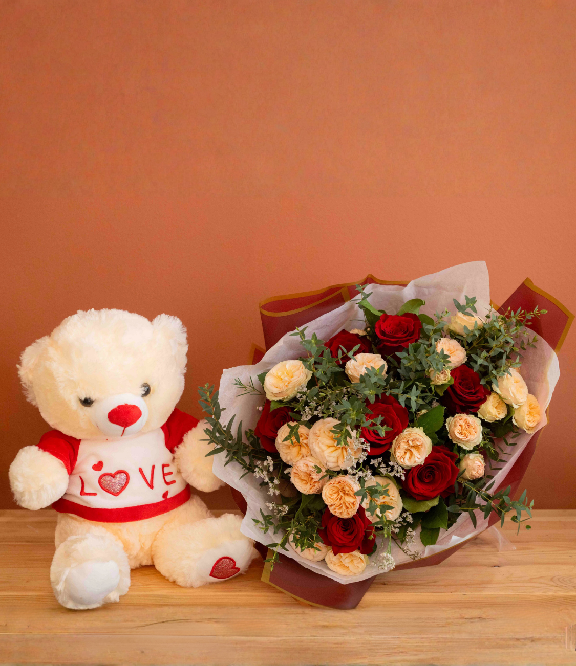Bouquet of red roses with green foliage in a clear glass vase tied with a raffia bow on a wooden surface against a terracotta background.
