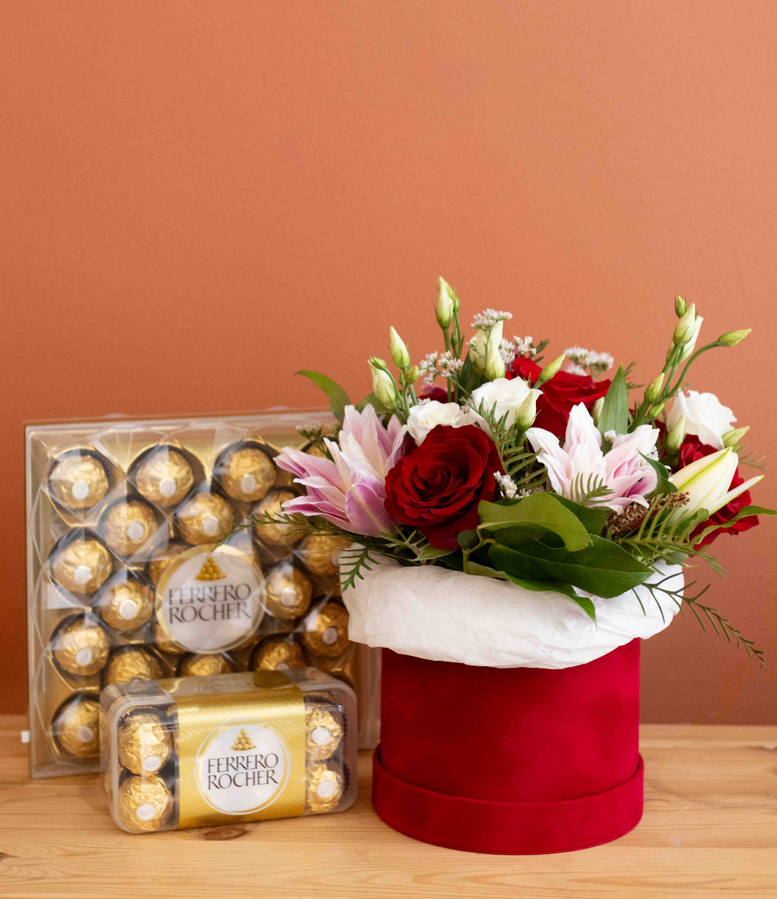 Bouquet of red roses with green foliage in a clear glass vase tied with a raffia bow on a wooden surface against a terracotta background.