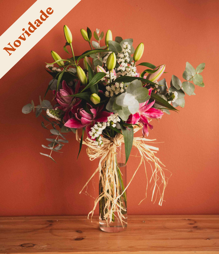 Bouquet of pink and peach flowers with green foliage tied with raffia in a clear glass vase on a wooden surface against a coral background.