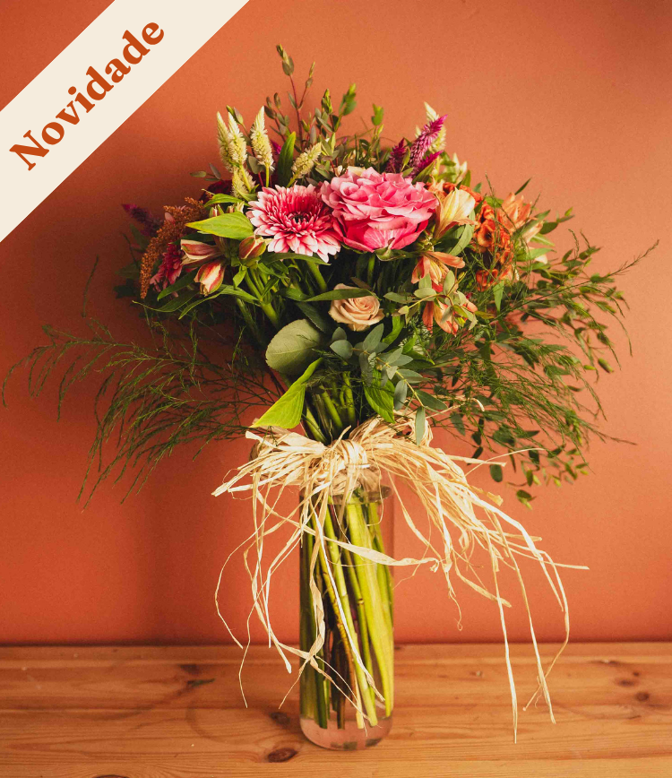Bouquet of pink and peach flowers with green foliage tied with raffia in a clear glass vase on a wooden surface against a coral background.