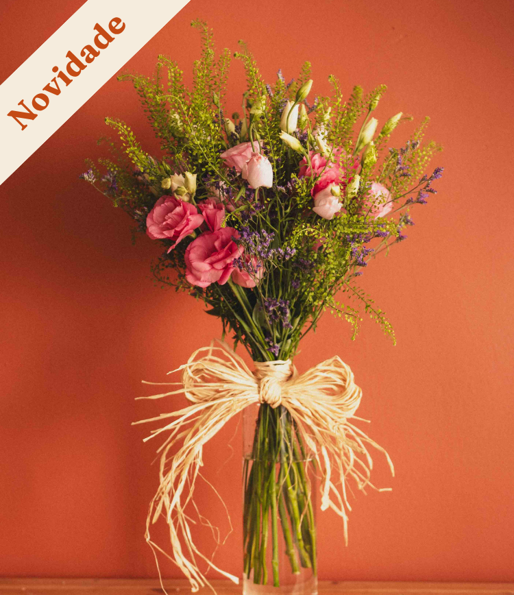Bouquet of pink and peach flowers with green foliage tied with raffia in a clear glass vase on a wooden surface against a coral background.