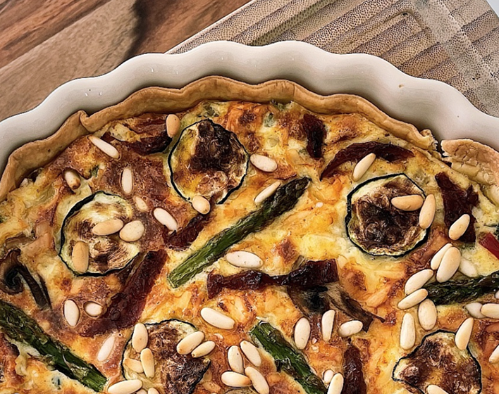 Close-up of a baked vegetable quiche topped with grilled zucchini slices, asparagus, sun-dried tomatoes, and pine nuts in a ceramic pie dish.