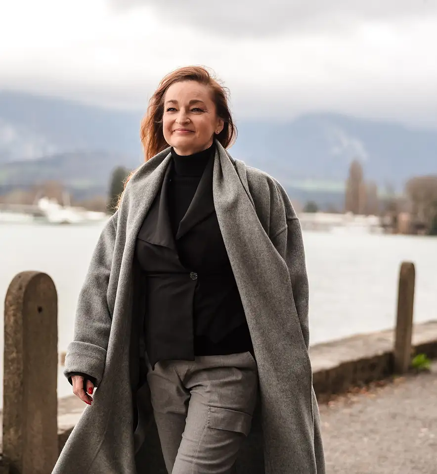 Woman with long hair wearing a grey coat and black top walking outdoors by a waterfront with mountains in the background.