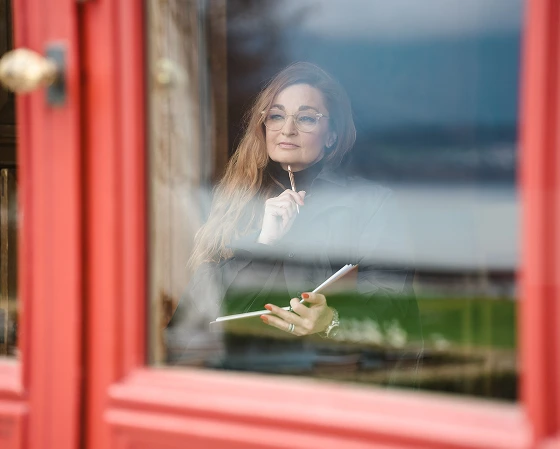 Woman with glasses holding a pencil and notebook, looking thoughtfully through a window with reflections.
