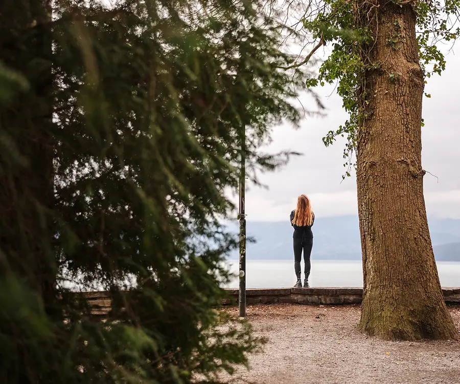 Person with long hair standing on a stone ledge, looking out at a lake surrounded by mountains, framed by trees.