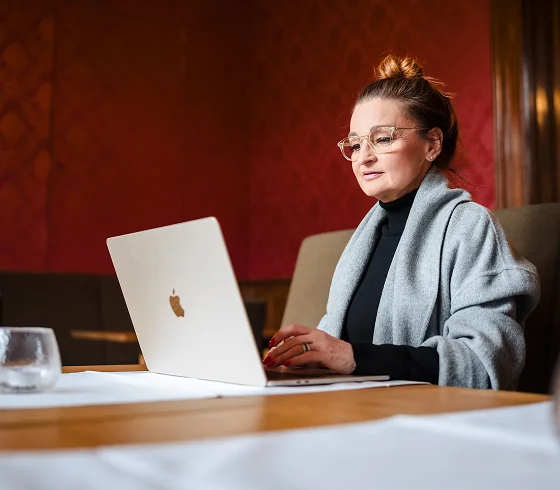 Woman with glasses and a gray shawl working on a silver laptop at a wooden table in a warmly lit room.