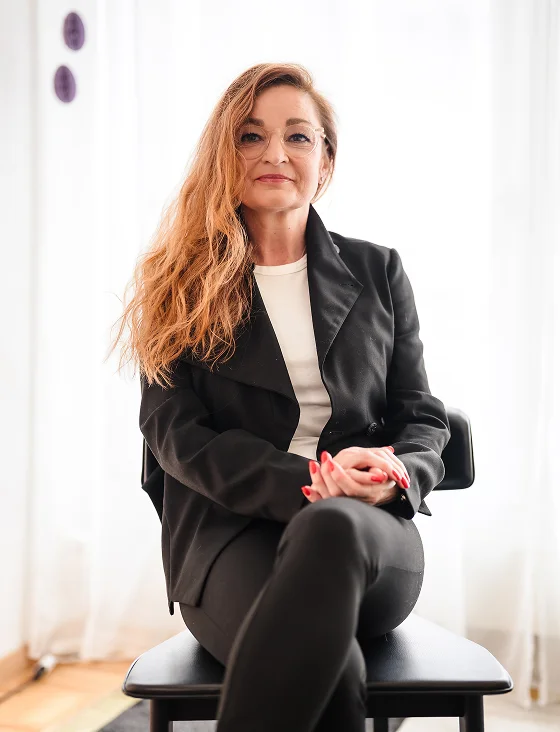 Confident woman with long wavy hair and glasses sitting cross-legged on a chair wearing a black jacket and white shirt.
