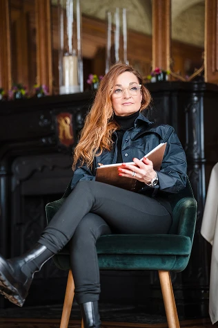 Woman with glasses and long red hair seated in a green velvet chair holding an open notebook in a room with dark wooden decor.