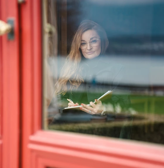 Woman with long hair and glasses writing in a notebook, seen through a window with reflections.