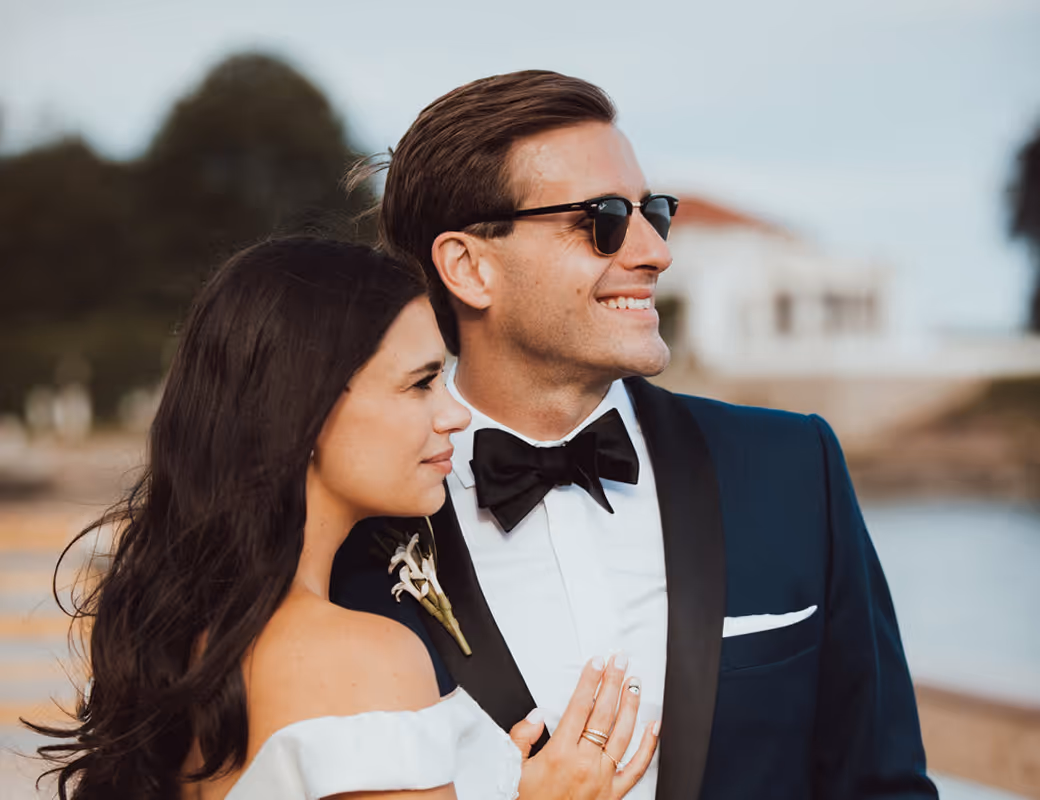 Couple dressed in wedding attire embracing outdoors, with the man wearing sunglasses and a tuxedo, and the woman in an off-shoulder white dress.