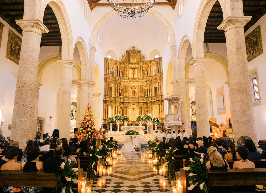 Interior of a church with tall stone arches and a decorated altar holding a wedding ceremony, with guests seated on wooden pews and candles lining the aisle.