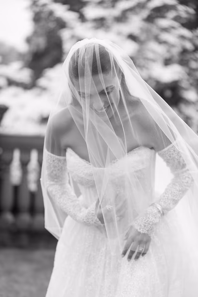 Bride in a lace wedding dress and veil looking down with a smile in a garden setting.