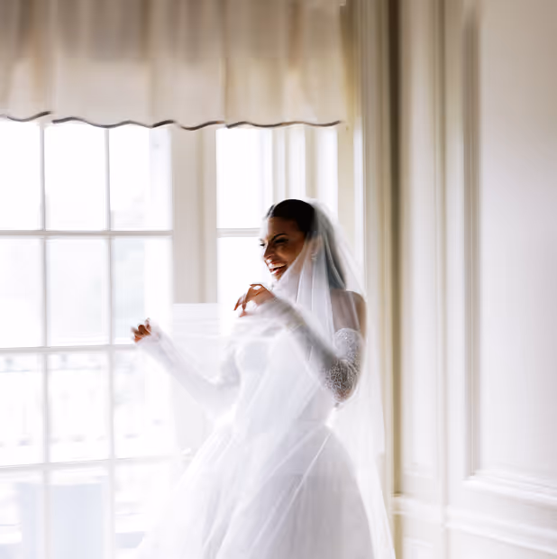 Smiling bride wearing a white wedding dress and veil standing in front of large bright windows.
