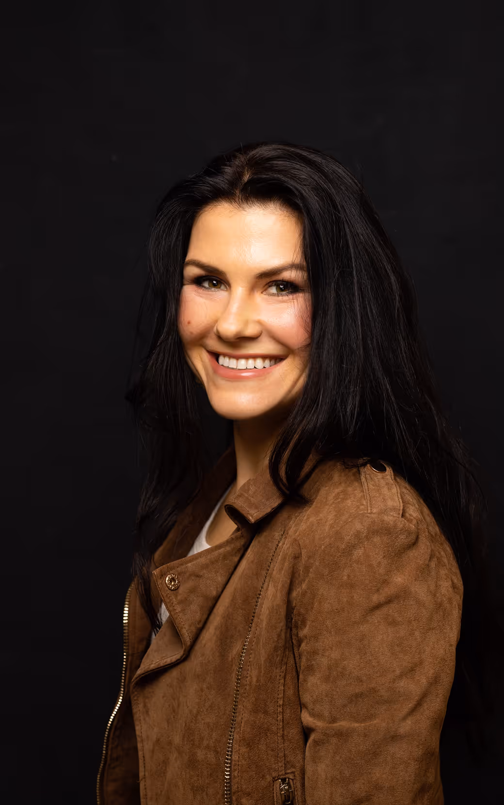 Smiling woman with long black hair wearing a brown suede jacket against a black background.