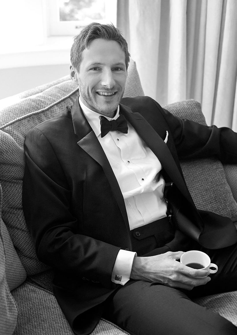 Smiling man in a tuxedo sitting on a couch holding a cup of coffee.