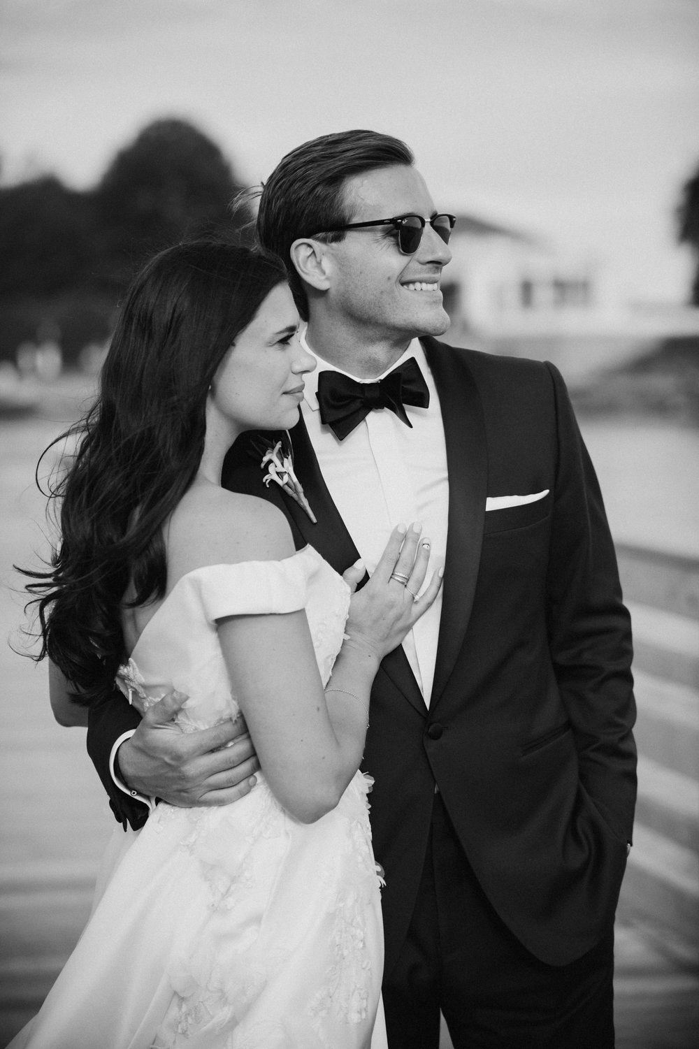 Black and white photo of a bride in an off-shoulder gown and a groom in a tuxedo with sunglasses embracing outdoors.