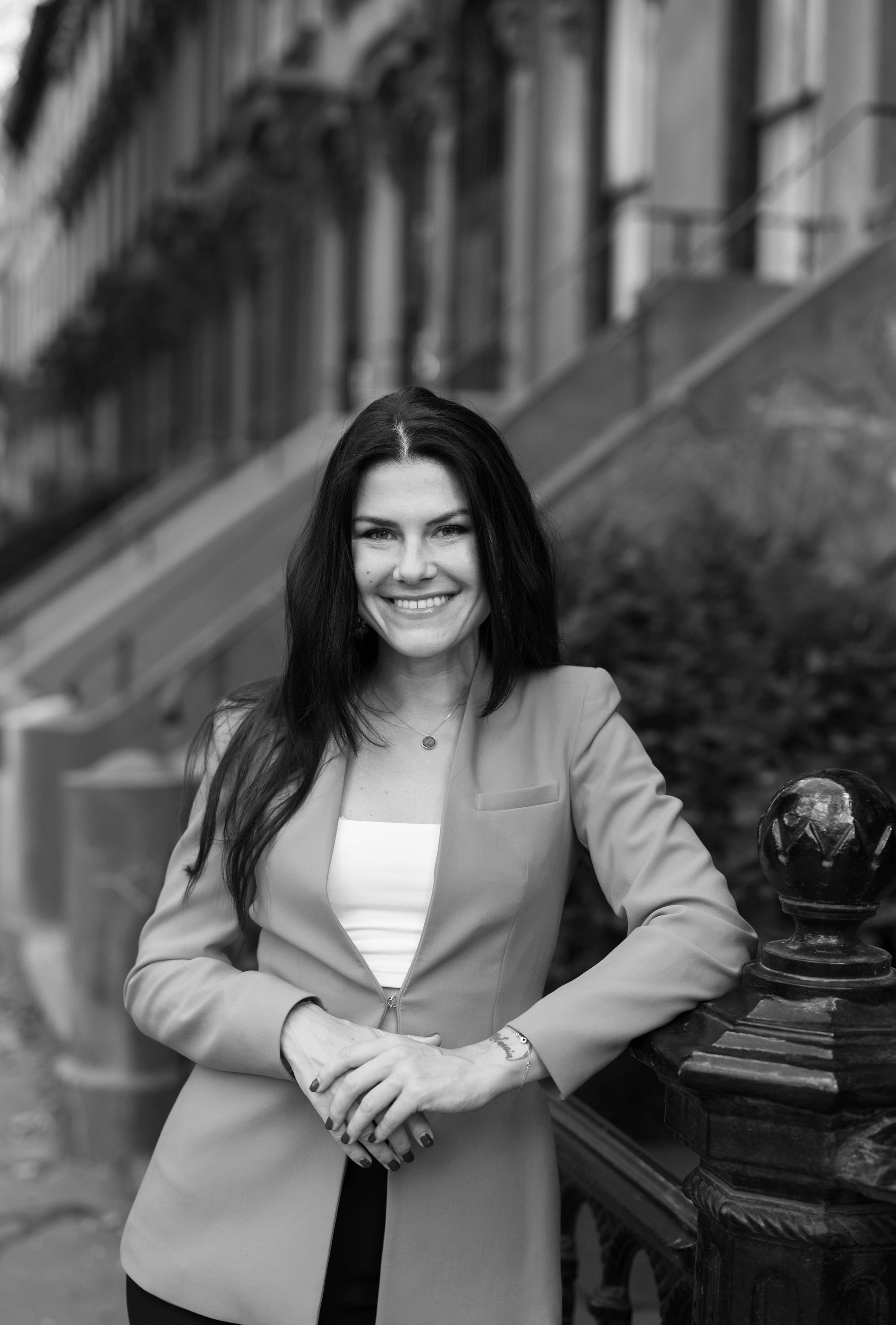 Smiling woman with long dark hair wearing an orange blazer and white top, leaning on a black railing with steps and greenery in the background.