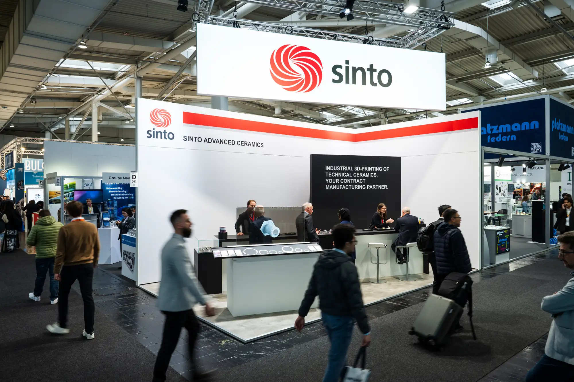 Sinto Advanced Ceramics booth at a trade show with people walking and talking inside the exhibit.