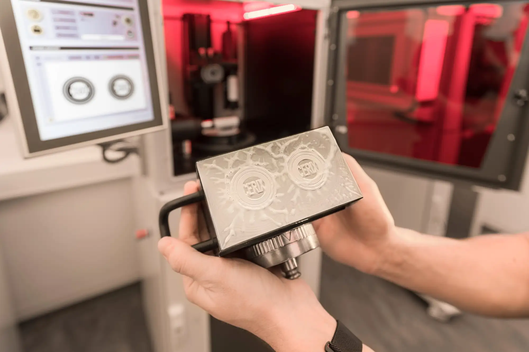 Person holding a 3d-printing building plattform with 'CERIX' embossed, near a computer and red-lit machinery.