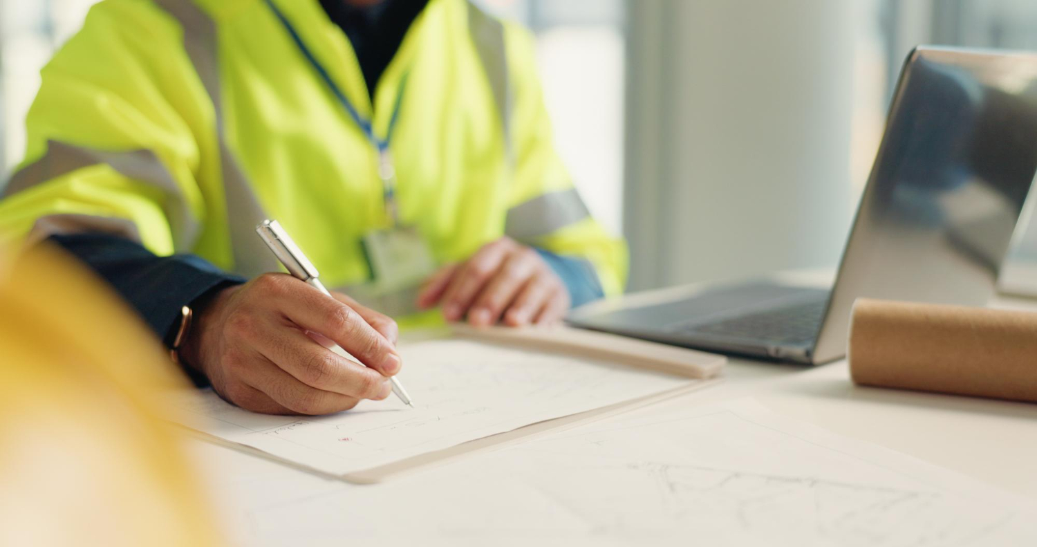 Henkilö keltaisessa heijastinliivissä piirtää tai kirjoittaa paperille toimistopöydällä lähellä kannettavaa tietokonetta ja piirustuksia.