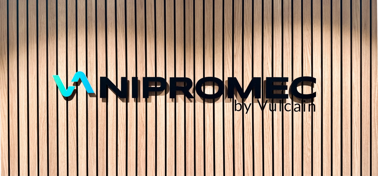 NIPROMEC-logo vaaleanpuunvärisellä pystysuuntaisella paneeliseinällä.
