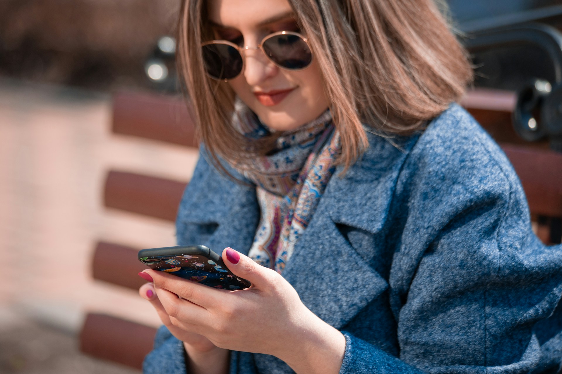 A woman in sunglasses and a blue coat sitting on a bench and looking at her smartphone.