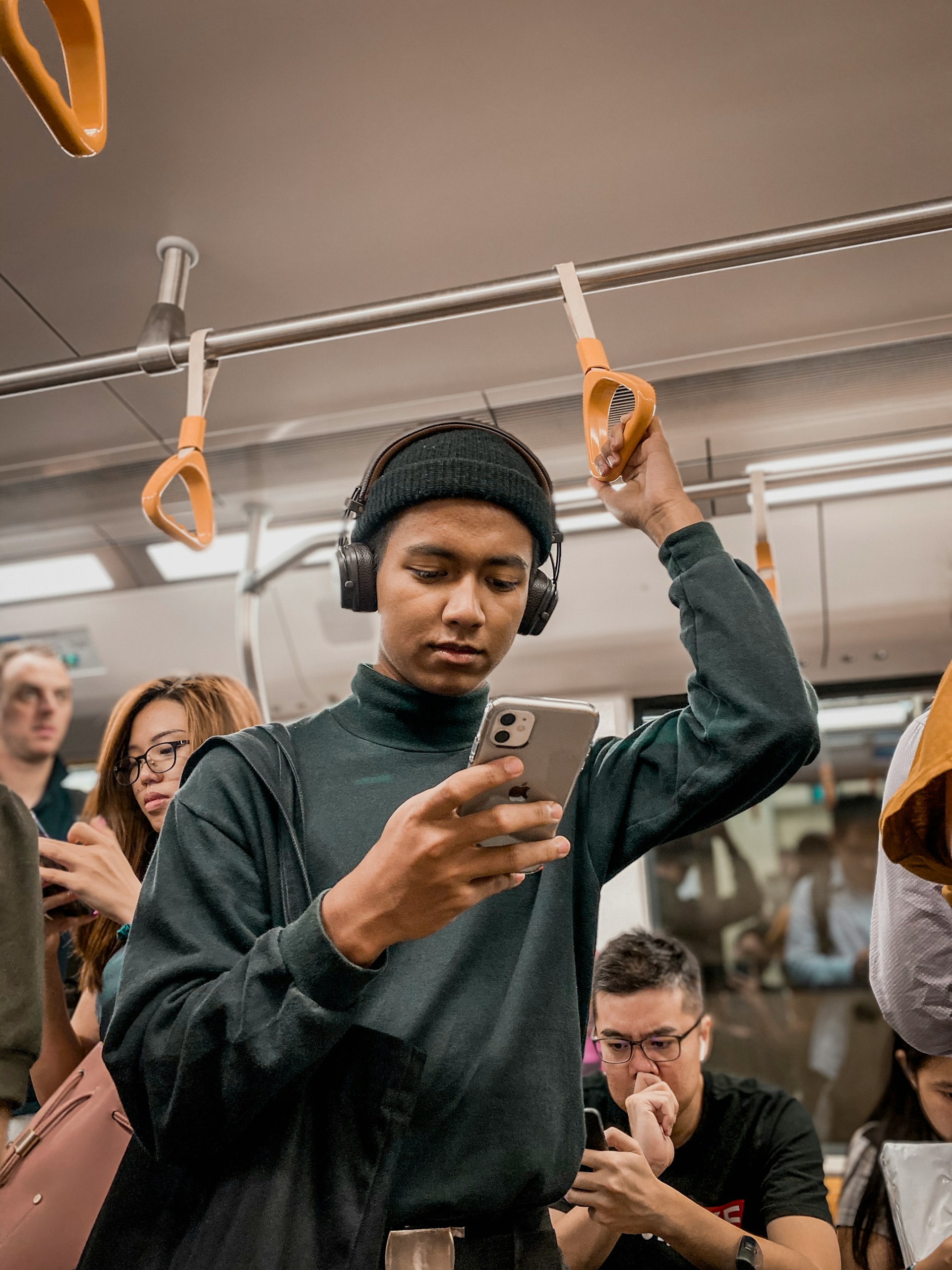 A young man with headphones and a black turtleneck is standing in a crowded subway or train car, holding onto a strap and looking at his smartphone.