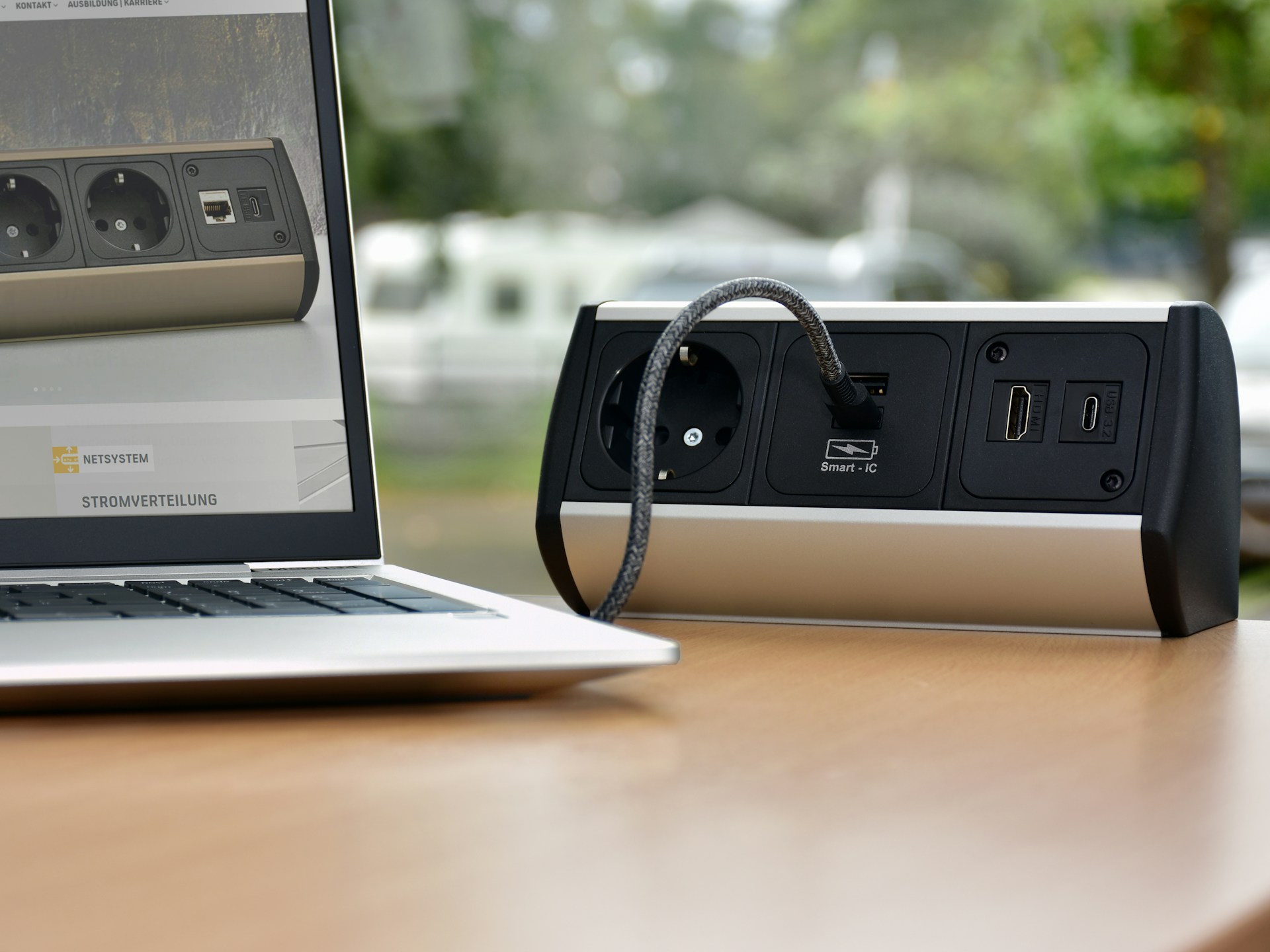 A silver and black multi-port power strip is sitting on a wooden table, with one power cord plugged in, positioned next to the screen of an open laptop.