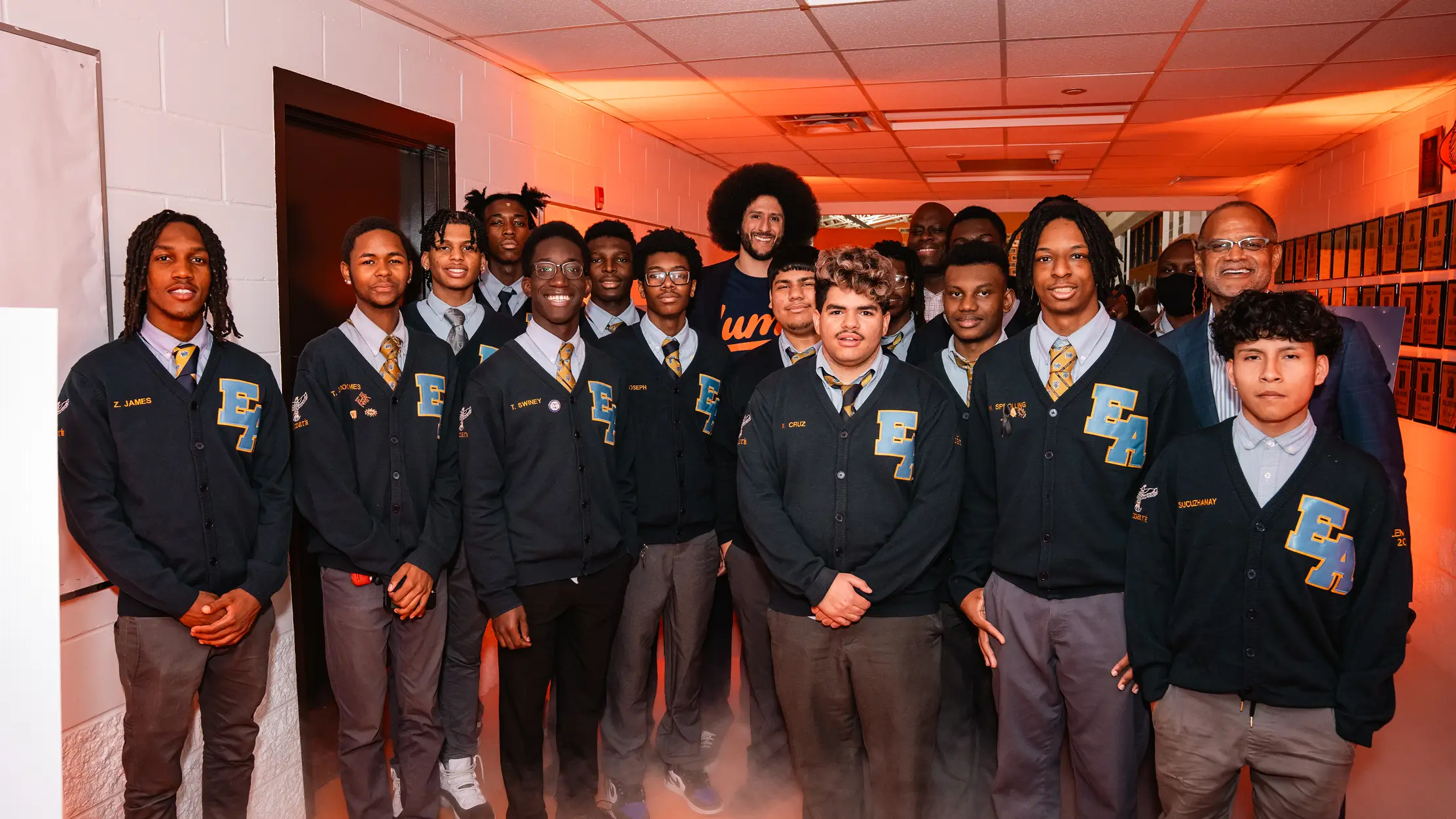 Group of young male students wearing matching black cardigans with EA initials and ties, posing indoors with two adult men.