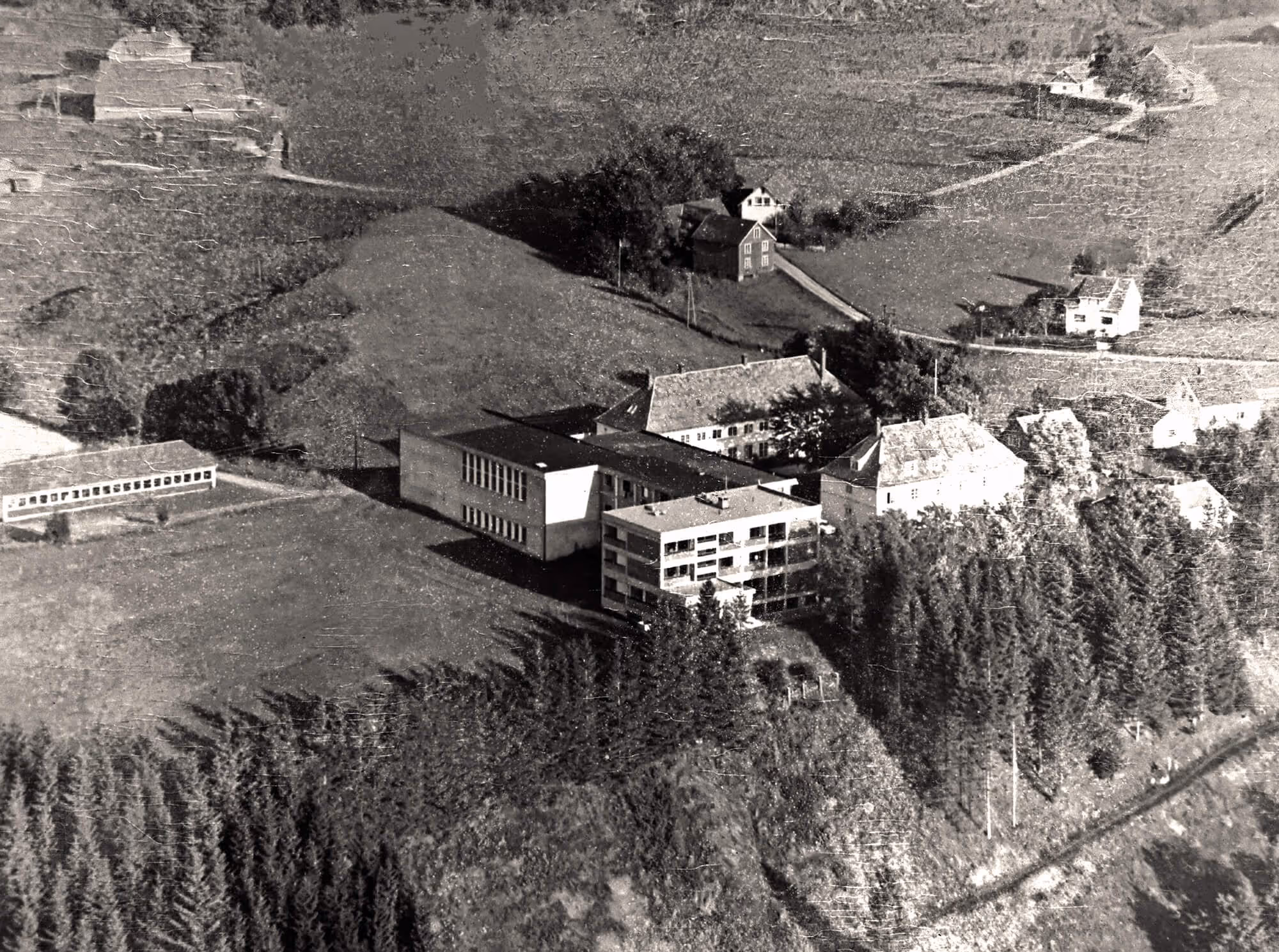 Luftfoto av et gårdsområde med flere bygninger, omgitt av skog og åpne jorder.