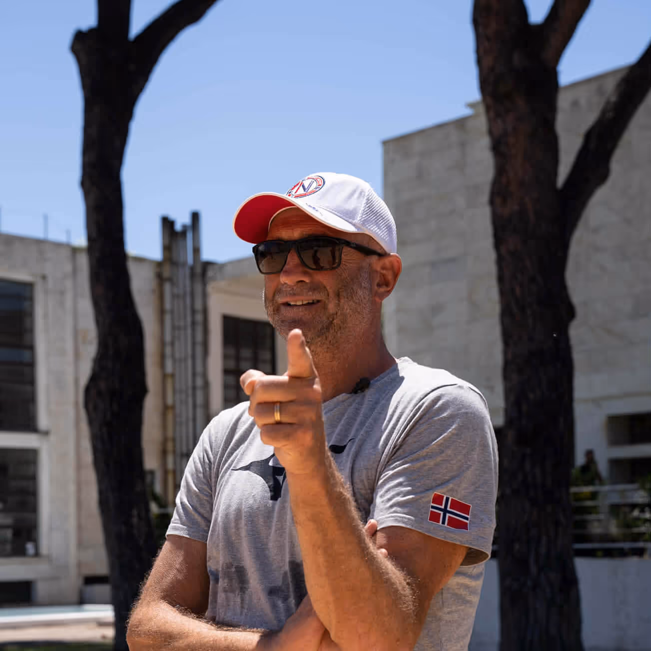 Mann i grå t-skjorte med norsk flagg på ermet, solbriller og hvit caps peker mot kamera utendørs.