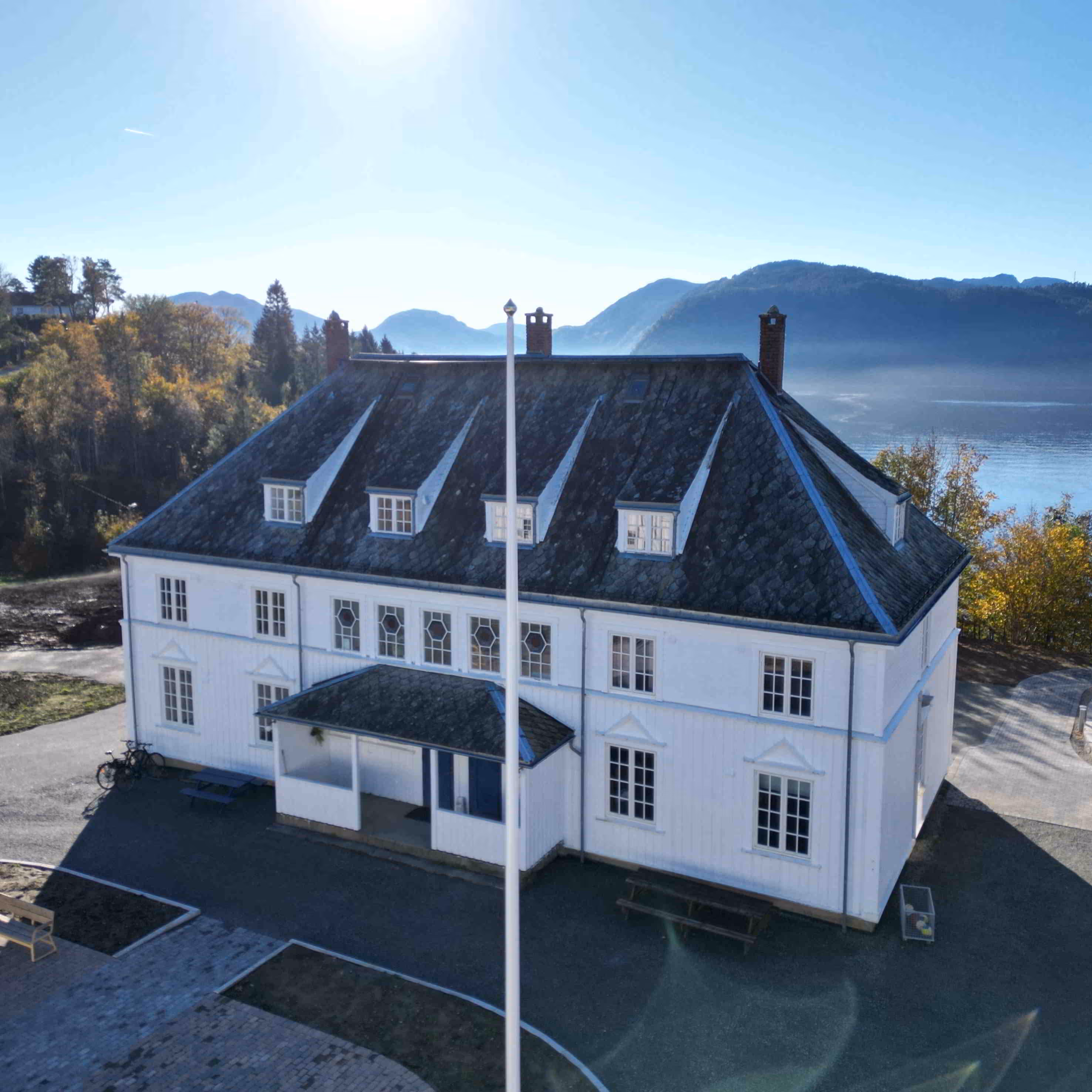 Hvitt to-etasjers hus med skrått tak og flere vinduer, omgitt av trær, fjell og vann under en blå himmel.